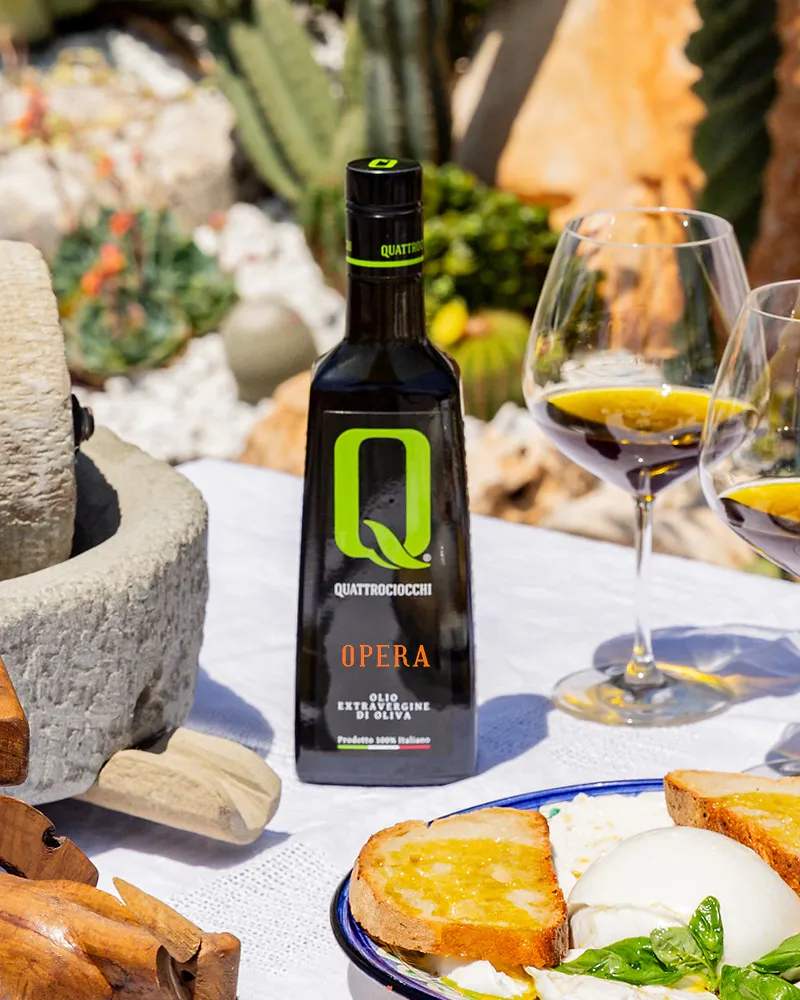 Flasche Quattrociocchi Olivenöl Extra Vergine 'Opera' neben zwei Gläsern mit Olivenöl und geröstetem Brot mit Mozzarella auf weißem Tischtuch im Freien